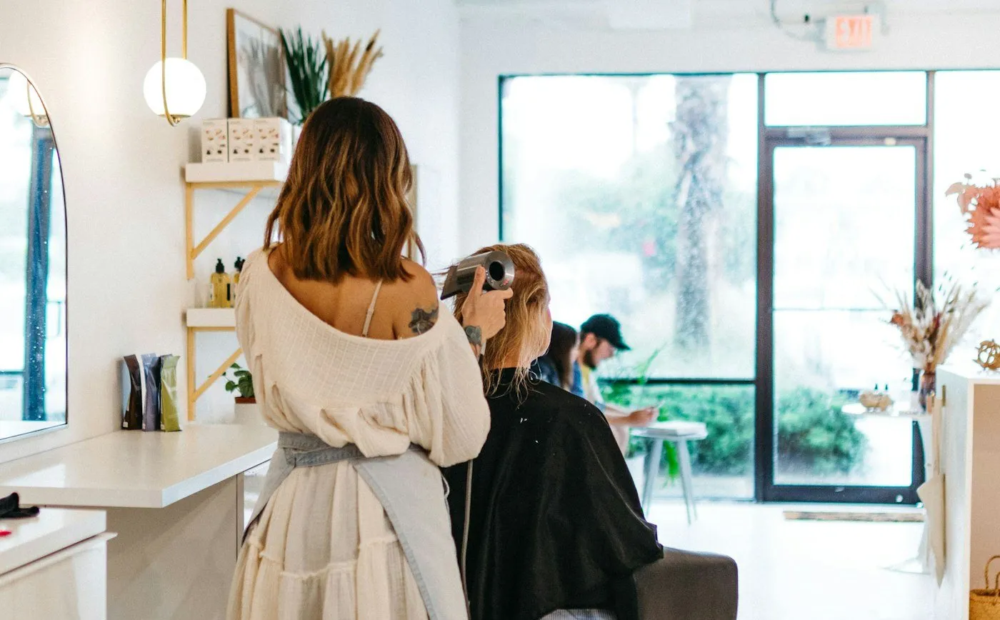 A stylist blow-drying a client’s hair in a bright salon with large windows